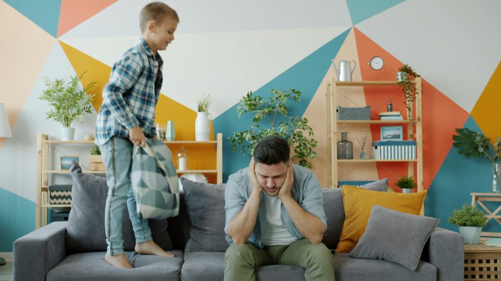 a child is jumping on a sofa next to a parent who seems frustrated and has their head in their hands